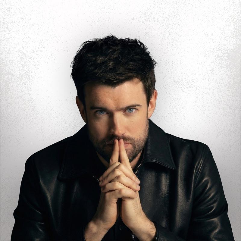 Jack Whitehall holding his hands to his lips looking pensively into the camera.