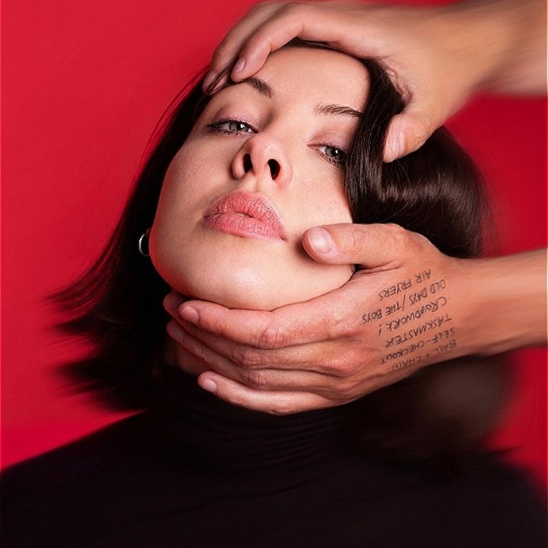 A blurry image of Harriet's face against a red background. Someone's hands reach in from out of frame and are holding the top of her head and her chin, pulling her head back so her face is tilted upwards