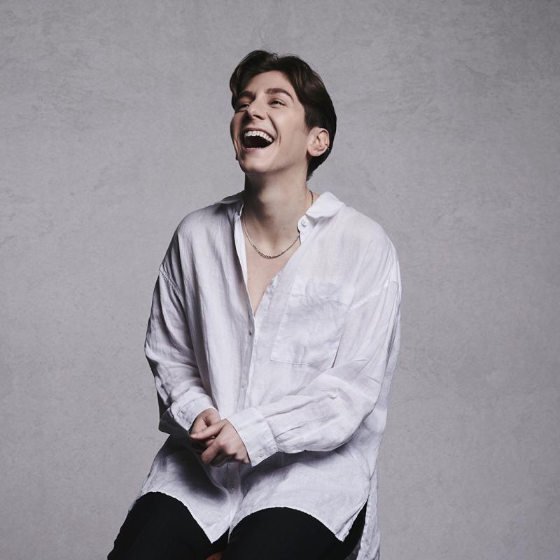 Sarah laughing on a stool wearing an oversized white shirt. There is a grey background.