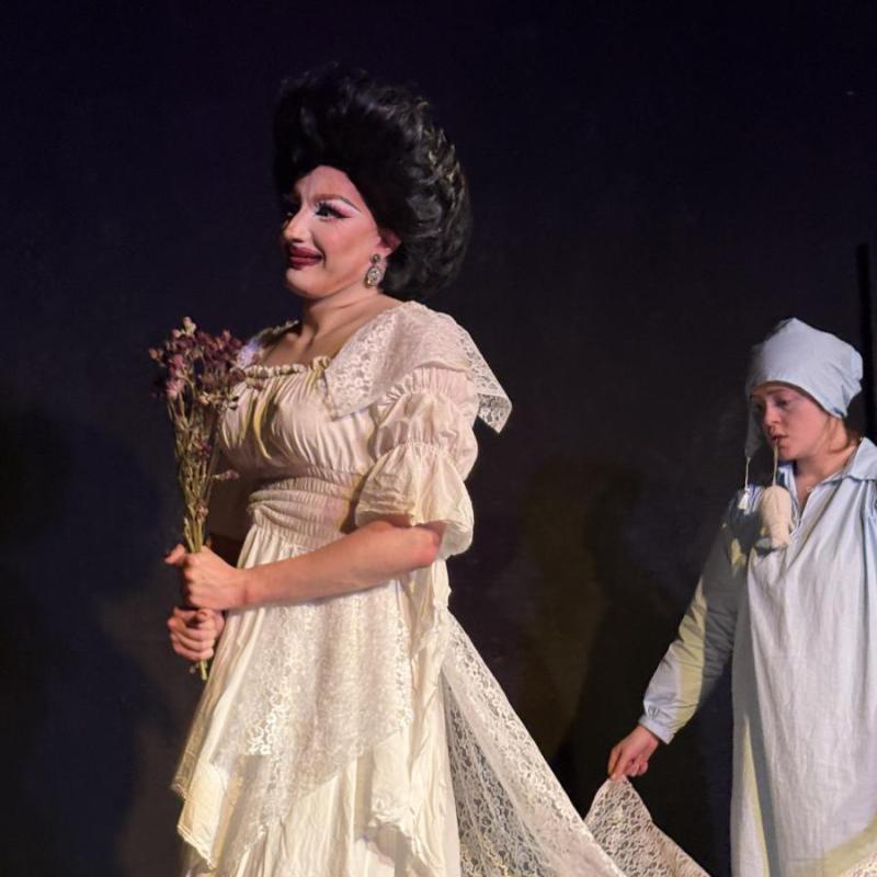 A victorian drag-queen wears a white wedding dress, clutches withered flowers and has the train of her dress held by her son Edvin, wearing powder blue night clothes and matching hat.