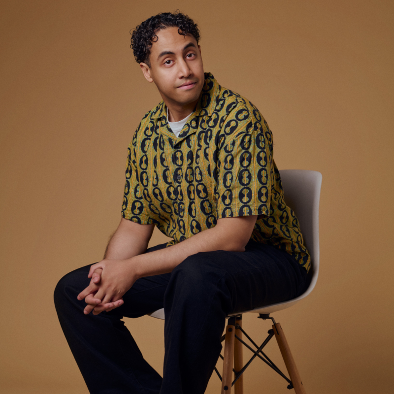 Omar sitting on a chair with his hands clasped gently in his lap. He wears a fun printed shirt against a beige brown background.