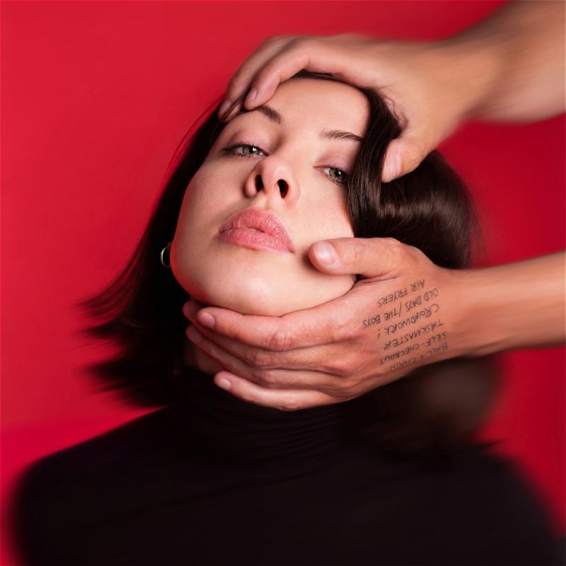 A blurry image of Harriet's face against a red background. Someone's hands reach in from out of frame and are holding the top of her head and her chin, pulling her head back so her face is tilted upwards