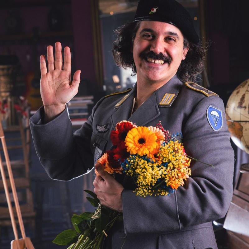 The performer wearing uniform and holding flowers waves at the camera with a large smile on their face.