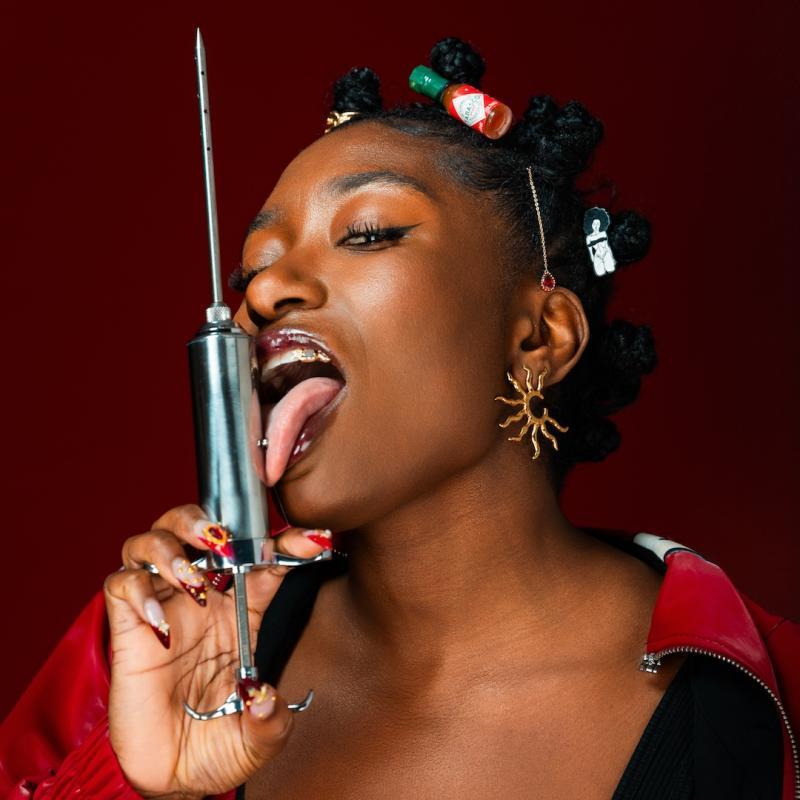 Sophie licks a large metallic syringe with an immense needle in a suggestive way. The syringe is an allegory for something else.
