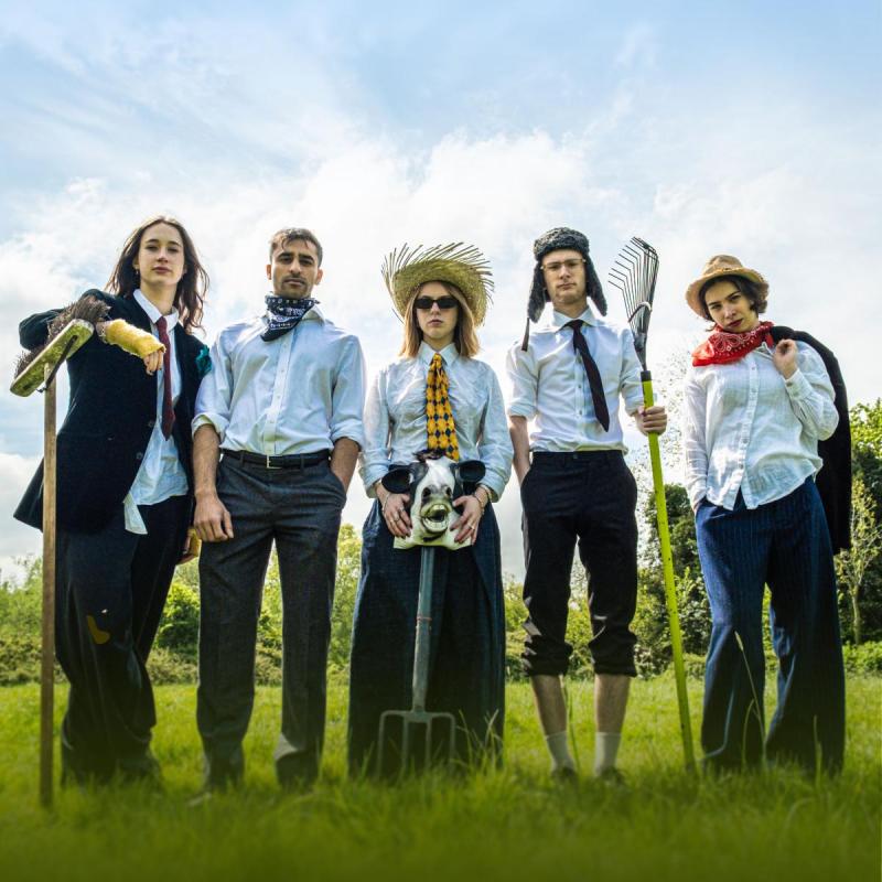 5 young adults in professional attire stand in a field holding farming equipment and looking at the camera.