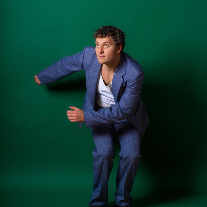 Jonathan wearing a blue blazer and trousers over a white t shirt crouches in front of a green backdrop. He is swinging his arms to one side and looking off camera to the left. 