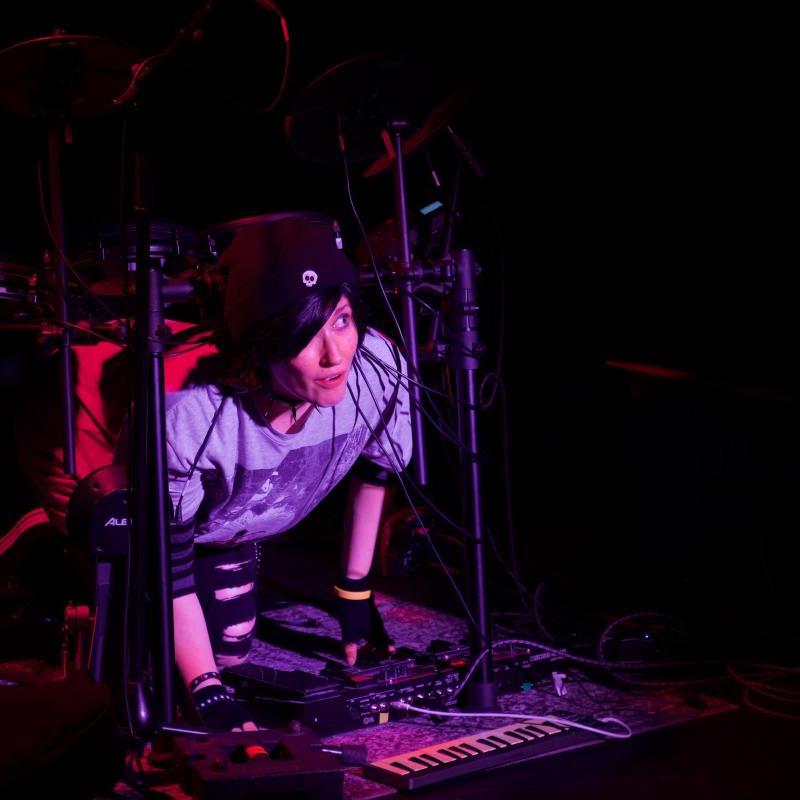 A woman in drag as an emo teenage boy kneels nervously under an electric drum kit, looking out at the audience.