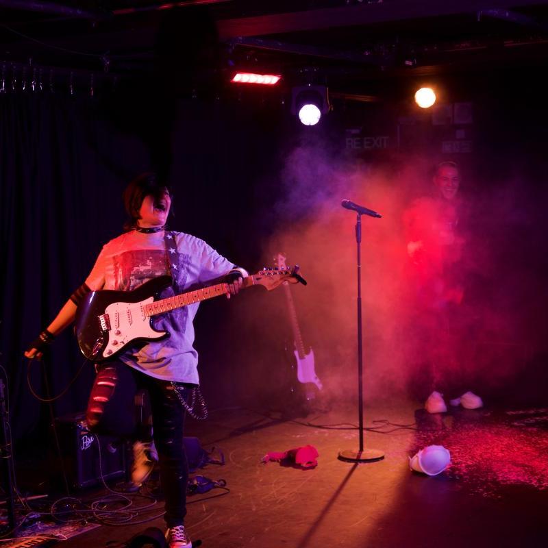 A woman in drag as an emo teenage boy holds her arms open, smiling, wearing an electric guitar as she is lit by coloured light and haze and a bra flies towards her.