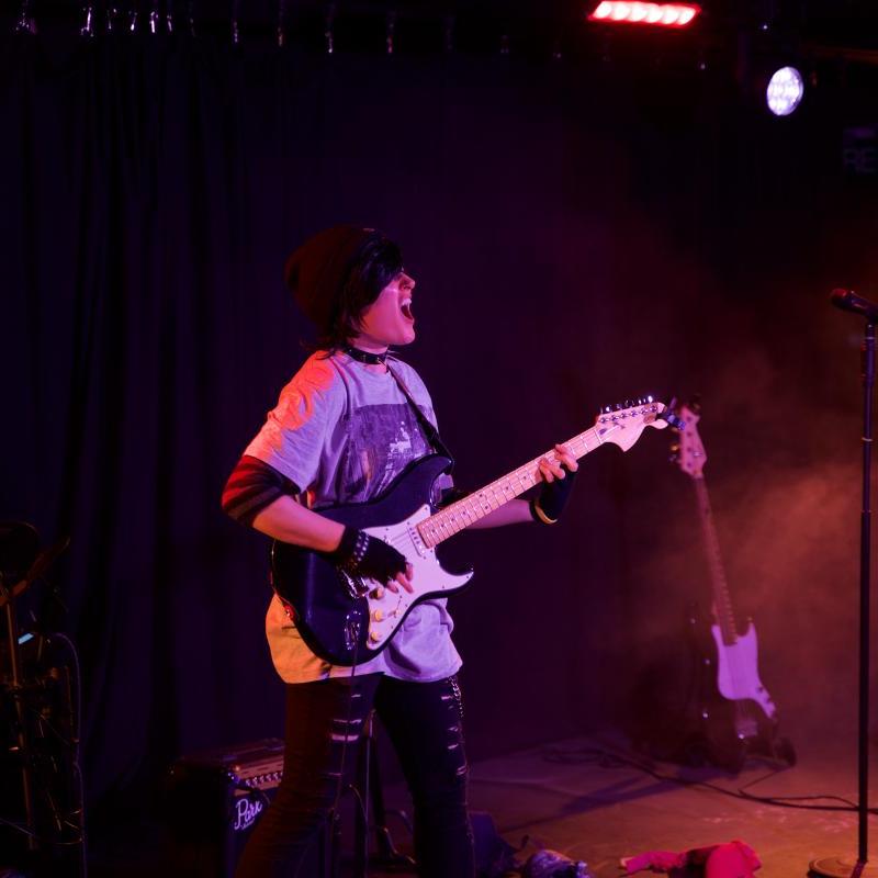 A woman in drag as an emo teenage boy plays the guitar and sings.