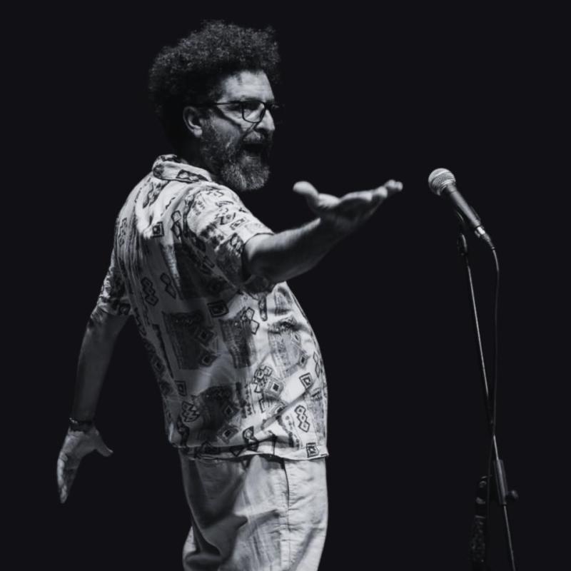 A black and white image of the performer standing in front of a microphone. They are reaching out their hand in a powerful pose. 