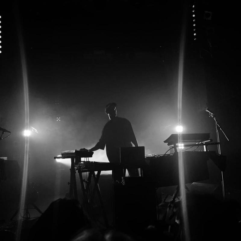 Isam stands in front of keyboards and other musical instruments, on a dark stage with bright stage lights