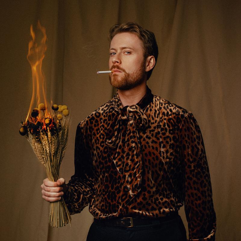 John wears a leopard print shirt and looks at the camera with a cigarette in their mouth. They are holding a bouquet of flowers that is on fire. 