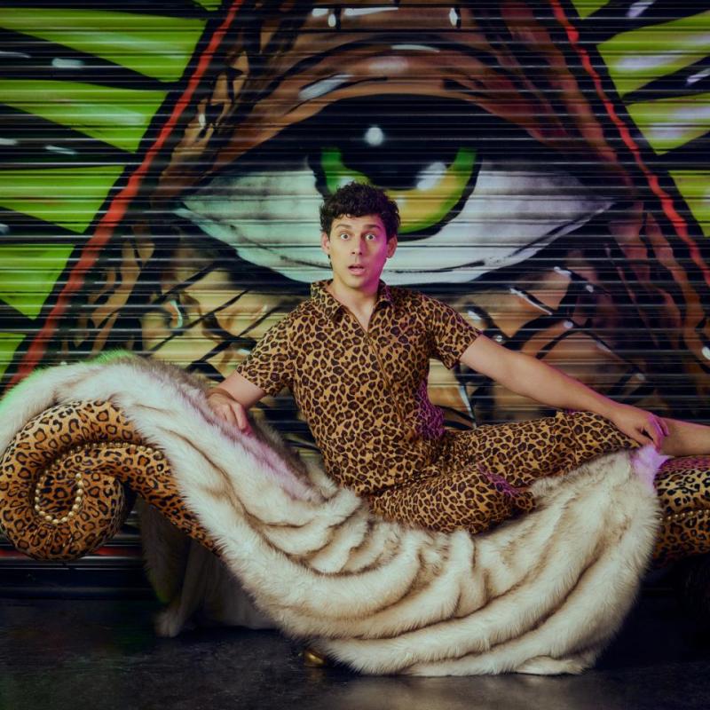 Matt wearing leopard print shirt and trousers, sat on a tiger print blanket on a leopard print chaise lounge. The chaise lounge is in front of a shutter graffiti-d with an eye in a triangle. 