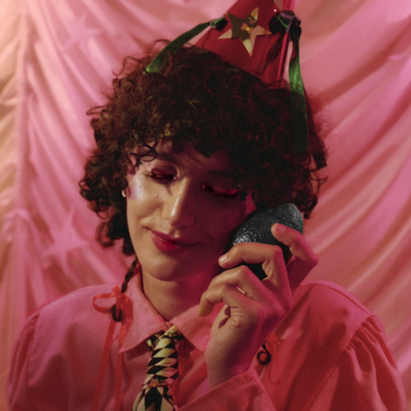The performer is dressed in a pink shirt, tie and party hat. They look at peace as they hold an avocado to their cheek.