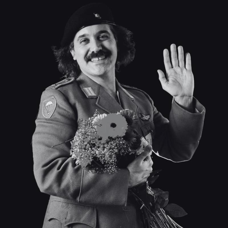 A black and white image of the performer wearing uniform and holding flowers waves at the camera with a large smile on their face.
