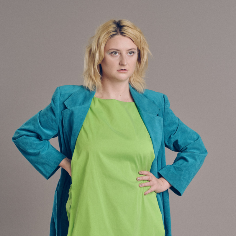 Lorna stands posing with their hands on their waist. They are wearing a green dress and a blue jacket and boots. 