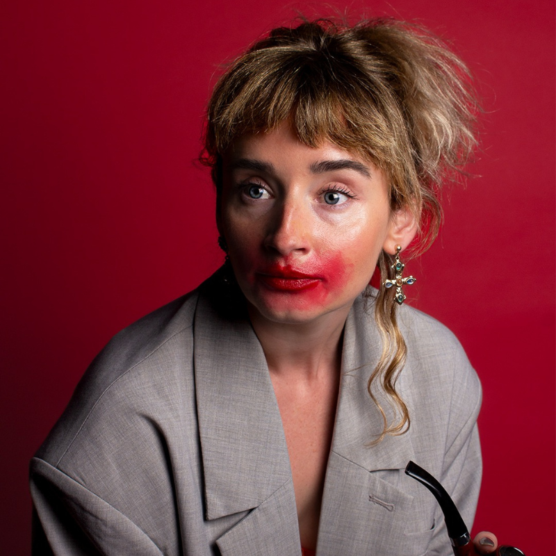 Molly is sitting against a red backdrop. She's wearing a grey blazer and holding a pipe. She has bright red lipstick smeared across her blank expression.