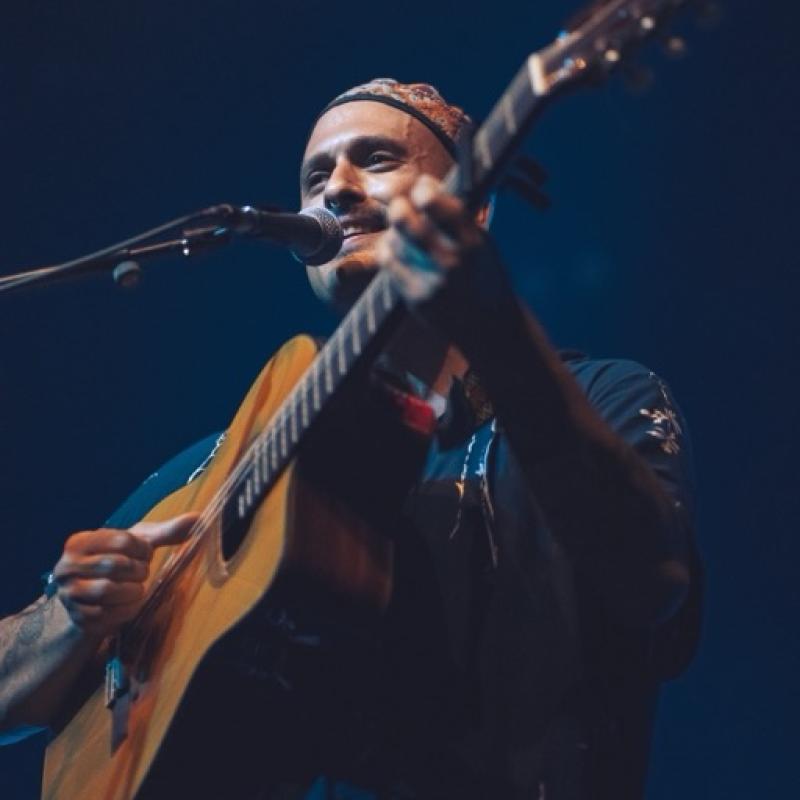 Ahmed stands behind a microphone on stage, playing a guitar