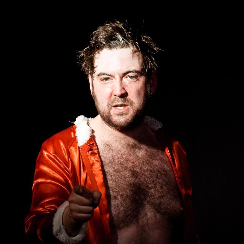Nick, wearing a christmassy red shirt with white fur trim, points at the camera against a black background