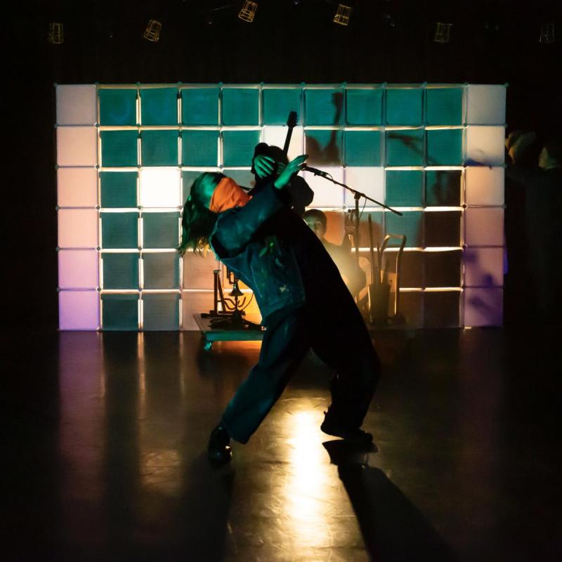The performer, dressed all in green, leans back whilst stood under a spotlight on stage.