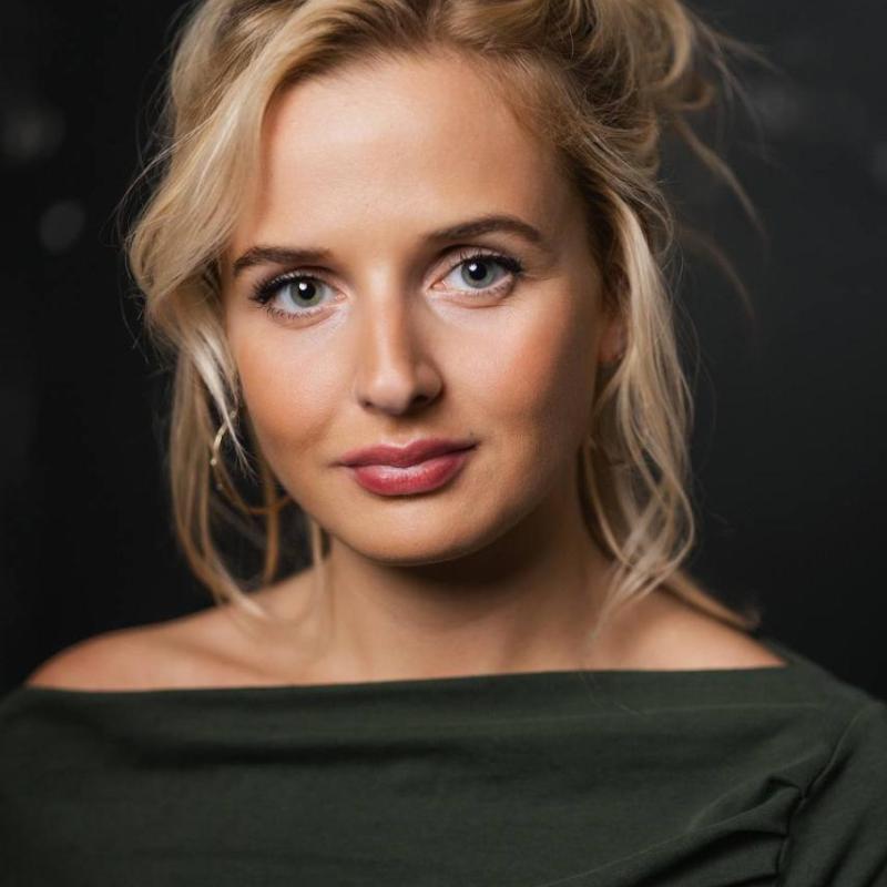 Headshot of Alana. Blonde hair, large gold hoops earrings and a slash neck grey top.