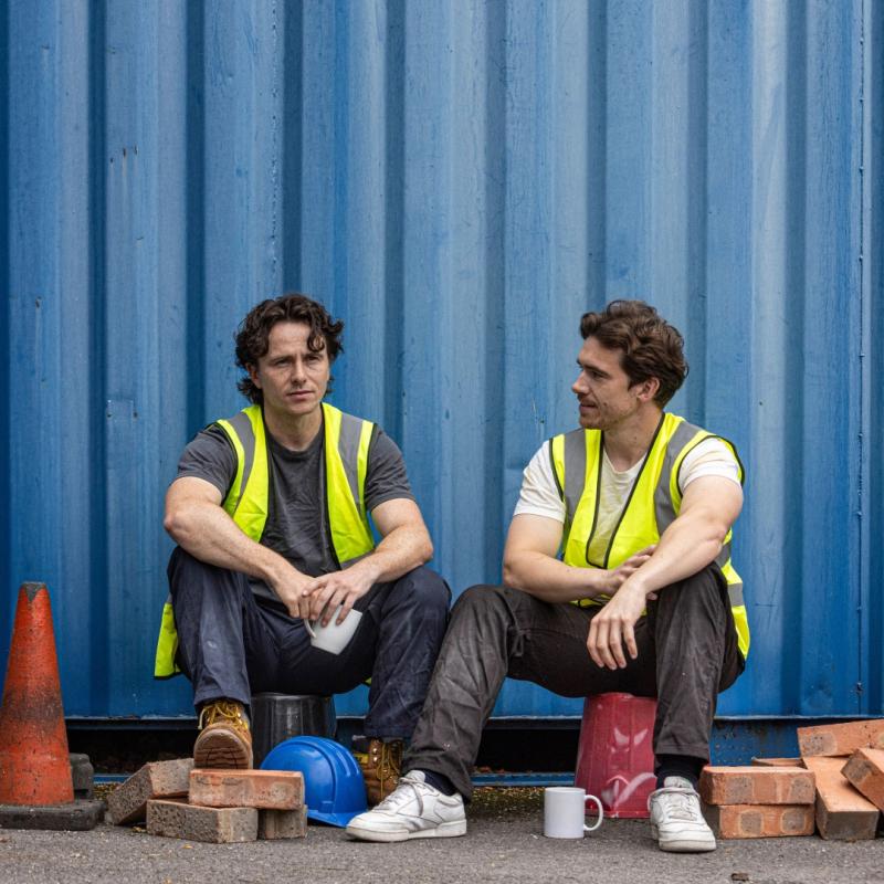 Two people are sat on buckets. They are both wearing high vis vests and are surrounded by building bricks.
