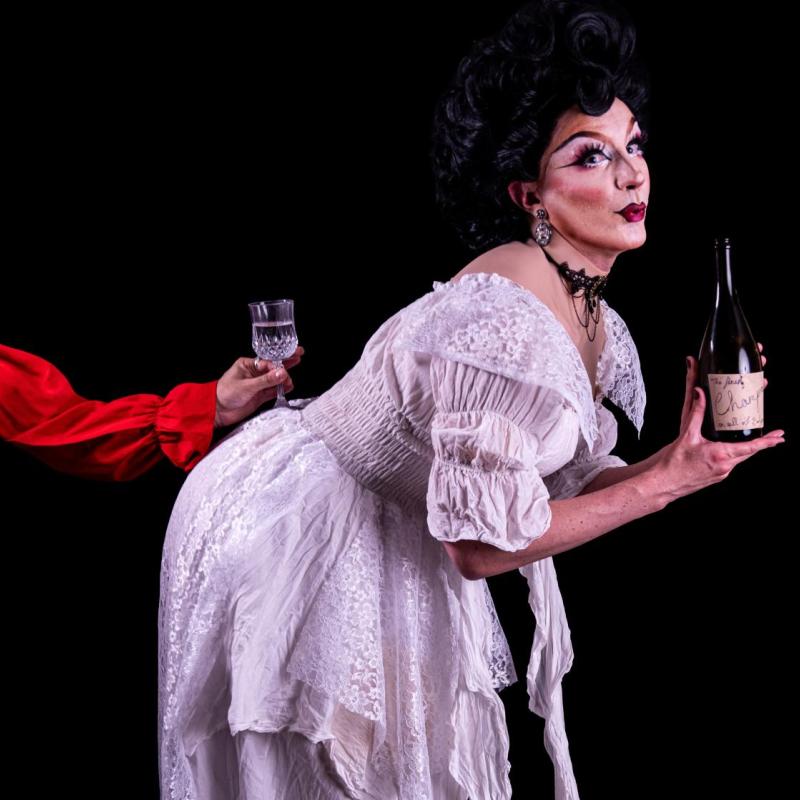 A drag queen in a white wedding dress leans forward, holding a champagne bottle, a red-sleeved hand balances a champagne glass on her back.
