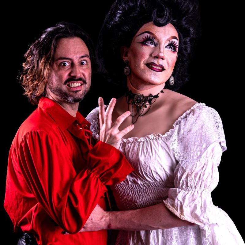 A drag queen in a white wedding dress and vampiric man embrace in a red victorian shirt.
