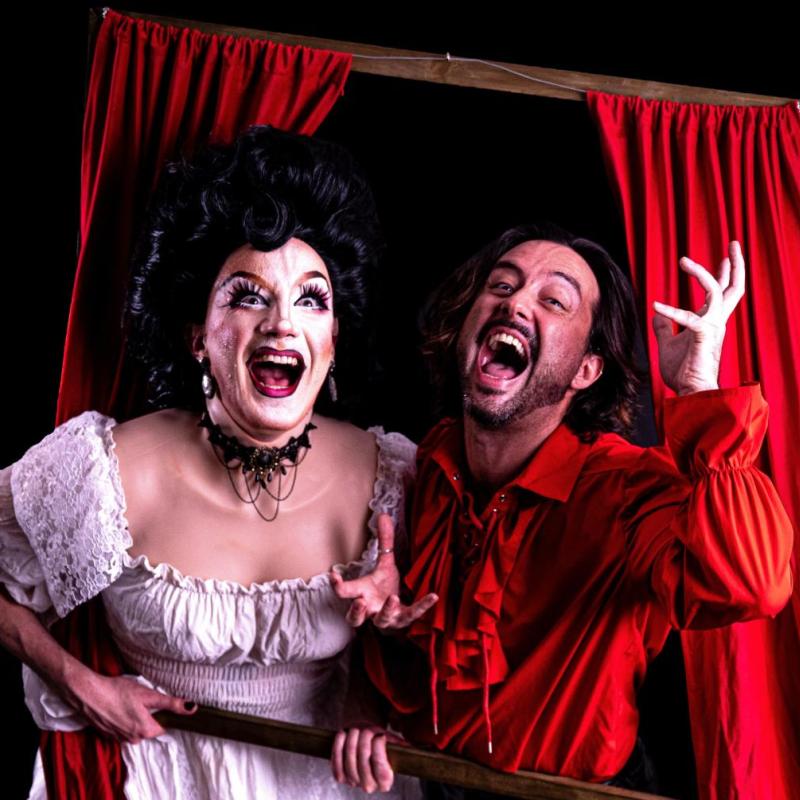 A drag queen in a white wedding dress and vampiric man in a red victorian shirt cackle out of a red curtained frame.