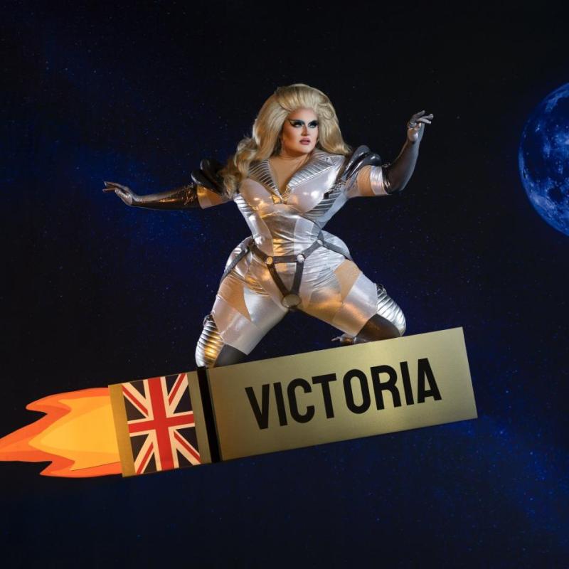 A person with blond hair wears a silver costume in space, with a planet behind them. They are kneeling on a rocket that has a union jack and says "Victoria"