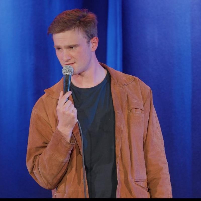 Josh Berry standing on stage holding a microphone against a blue curtain