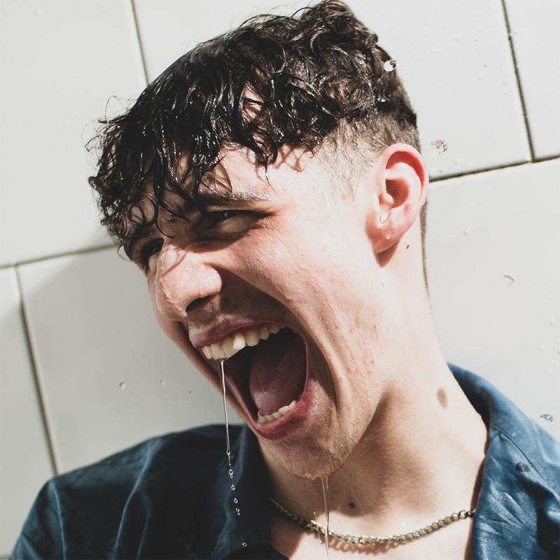 A man with curly hair against a tiled background. His hair is wet and water is dripping from his face.