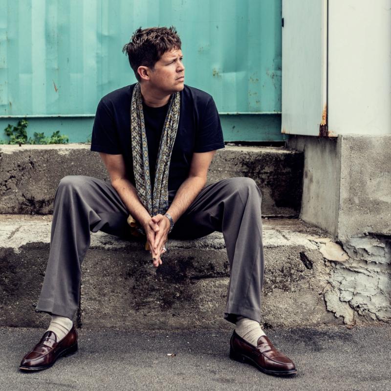 The performer sits on a concrete step wearing black and grey, looking into the distance