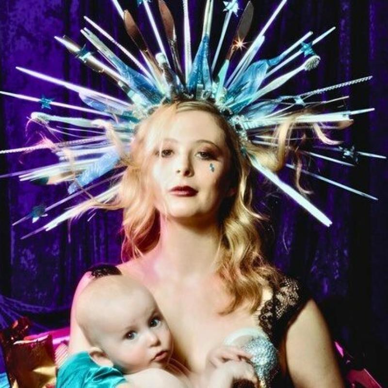 A woman holding a baby with wavy blonde hair is wearing a spiked crown