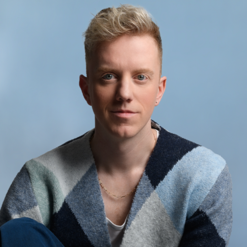 An image of James Barr, looking directly at the camera. They are wearing a blue tone block cardigan and sitting against a blue background