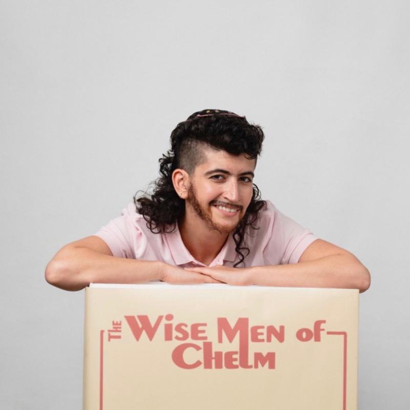 The performer smiles while leaning of a cardboard box which has has 'The Wise Men if Chelm' written across it in bold red lettering.